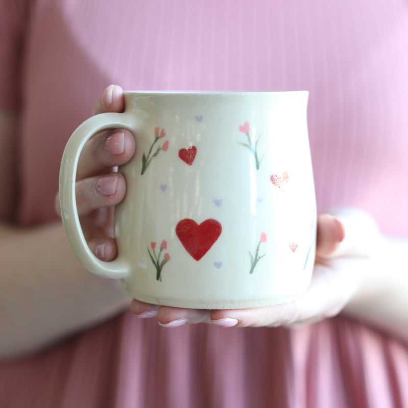 Person holding a mug with heart and flower designs against a pink background