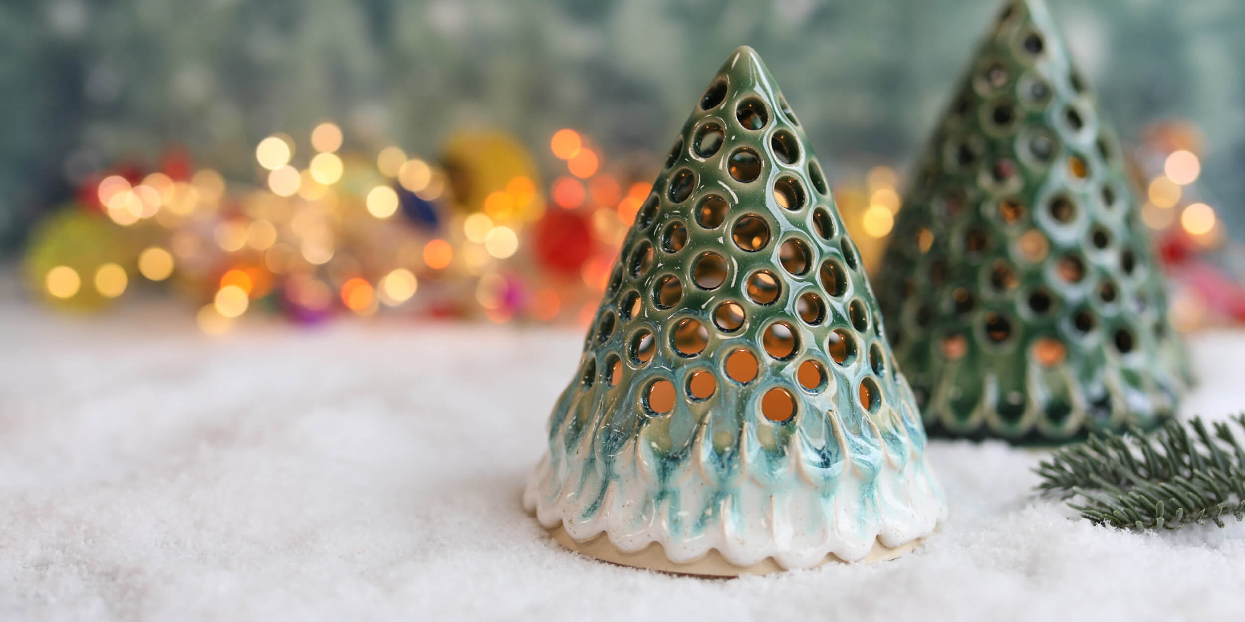 Two decorative ceramic Christmas tree ornaments with green and white glaze, featuring circular cutouts that reveal warm light, set in white snow with festive blurred lights in the background.