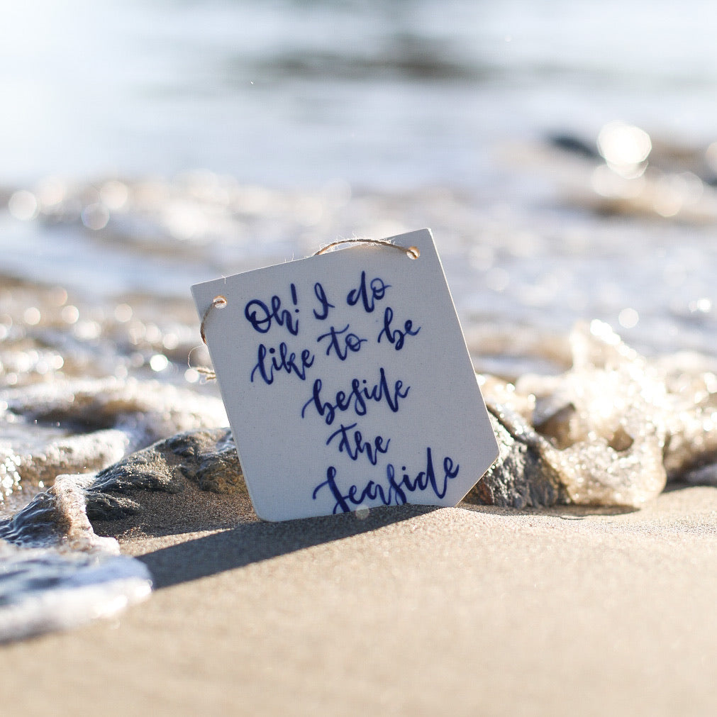 A ceramic banner with the quote ‘oh I do like to be beside the seaside’ in navy calligraphy.